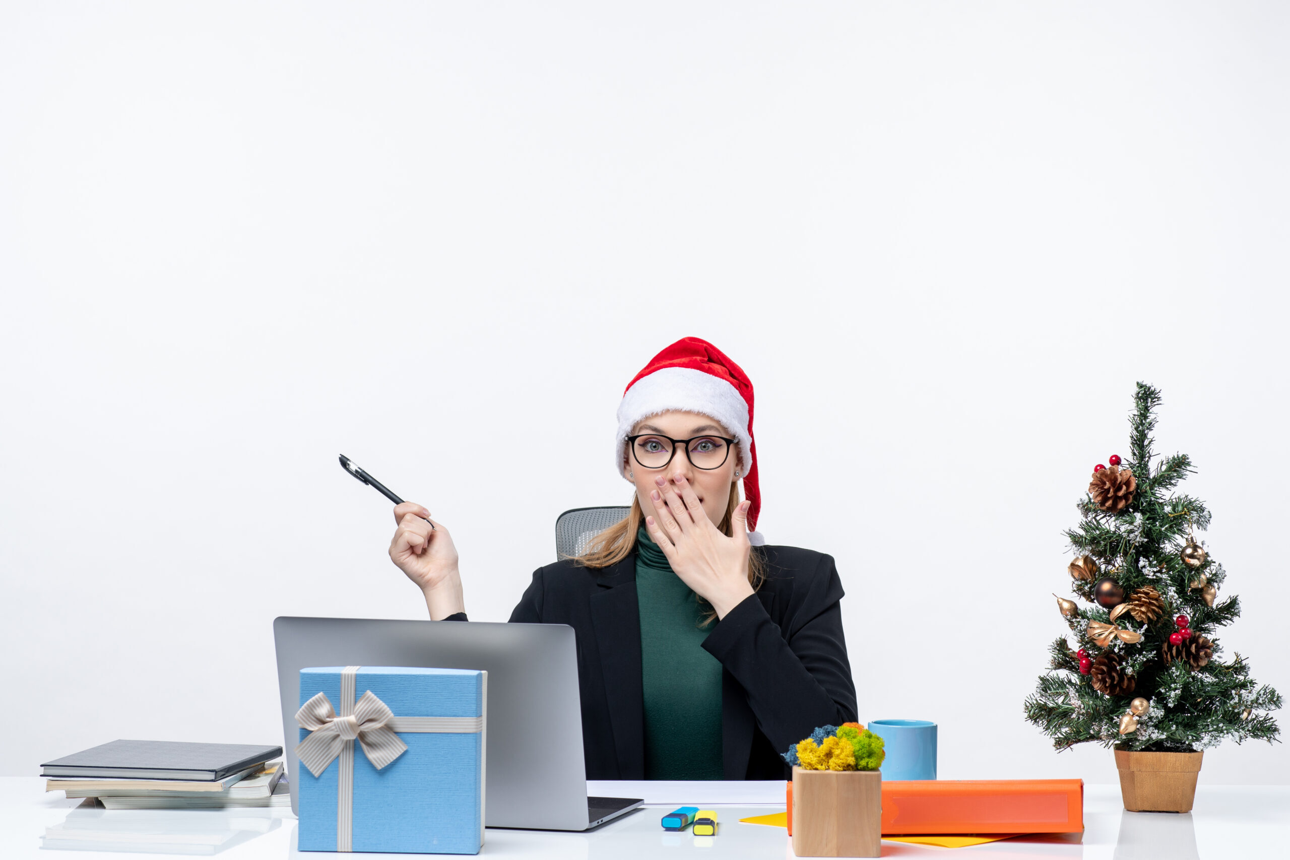 surprised-blonde-woman-with-santa-claus-hat-sitting-table-with-christmas-tree-gift-it-white-background